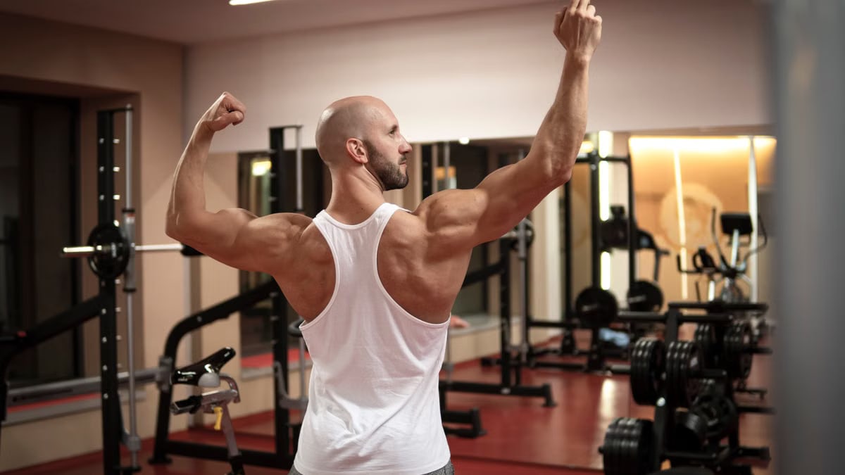 A muscular man in a gym flexes his back muscles wearing a white tank top.